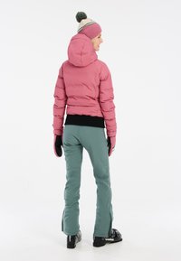 Rosa Daunenjacke mit Kapuze, schwarzen Bündchen und grünen Skihosen. Model trägt schwarze Skischuhe und eine gestrickte Mütze in Pink und Creme.
