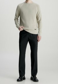 Beige Waffelpullover mit gerippten Bündchen, kombiniert mit schwarzen geraden Hosen und schwarzen Schnürschuhen. Minimalistisches Design, ohne Muster.