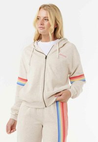 Beige zip-up hoodie met een voorkant rits, verstelbare capuchon en regenbooglijn accent op de mouwen. Zachte textuur.