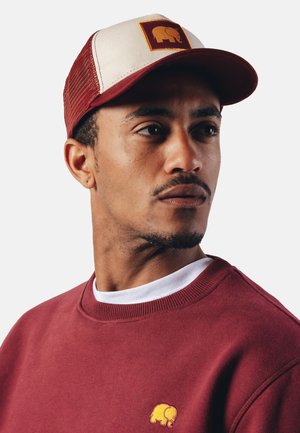 Casquette de camionneur avec un devant maroon, un dos en maille crème et un patch carré représentant un éléphant jaune. Sweat à capuche bordeaux avec un petit logo jaune.