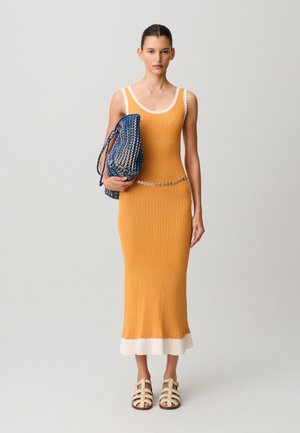 Mujer con un vestido midi sin mangas color mostaza con ribetes blancos, sandalias beige, un cinturón de cadena plateada y sosteniendo un bolso tejido azul y blanco.