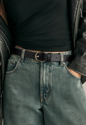 Cintura nera in pelle con fibbia dorata indossata sopra jeans verde chiaro con una trama sbiadita. In vista un top scuro a coste e una giacca di pelle.