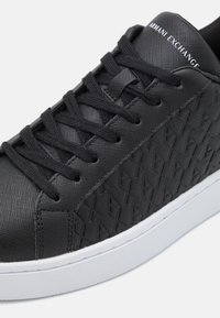 Sapatilha preta feita de tecido texturizado com um padrão tecido, apresentando atacadores planos pretos e uma sola de borracha branca. Logótipo "Armani Exchange" na língua.