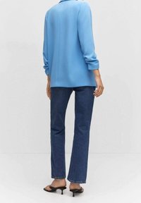 Sininen pitkähihainen rento paita, yhdistetty tummansinisiin suoralahkeisiin farkkuihin ja mustiin korollisiin sandaaleihin, jotka korostavat sileää tekstuuria.