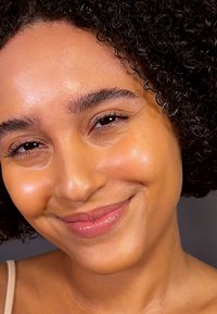 Närbild av en person med lockigt hår och en slät, lysande hy. Funktionerna inkluderar fylliga läppar och lätt makeup, mot en neutral bakgrund.