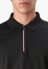 Polo negro con cuello texturizado y cierre en el cuello. Presenta una raya vertical en rojo, blanco y azul en el cierre.