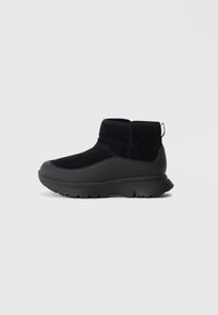 Botas de tornozelo pretas feitas de camurça com sola em borracha. Apresentam um cabedal liso com bico arredondado e uma alça na parte de trás para facilitar o calce.