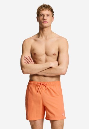 Hemdloser Mann mit lockigem blondem Haar trägt orange Badehosen und steht mit verschränkten Armen vor einem schlichten weißen Hintergrund.