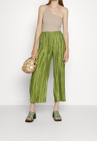 Zielone plisowane spodnie o szerokich nogawkach, beżowy top na jedno ramię, zielone sandały na szerokim obcasie oraz pleciona torba o zaokrąglonym kształcie.