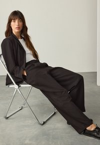 Blazer noir sur mesure et pantalon large associés à un t-shirt blanc. Chaussures en cuir noir. Modèle assis sur une chaise pliante métallique.