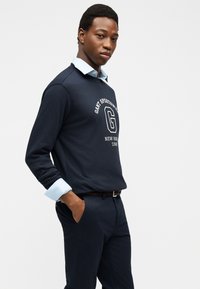 Hombre vestido con sudadera azul marino con logo blanco y camisa azul claro con cuello debajo, de pie con las manos en los bolsillos.