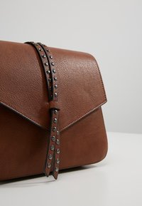 Brązowa skórzana torebka na ramię z klapką zapinaną na zatrzask, ozdobiona ćwiekami wzdłuż paska zapinającego oraz z teksturowanym wykończeniem.