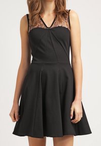 Robe noire en A avec un corsage ajusté, panneau en dentelle au niveau du col et jupe plissée. Présente un design sans manches et une encolure en V.