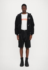 Black zip-up hoodie with contrasting white stitching, white t-shirt with colorful graphic, black shorts, and chunky black boots.