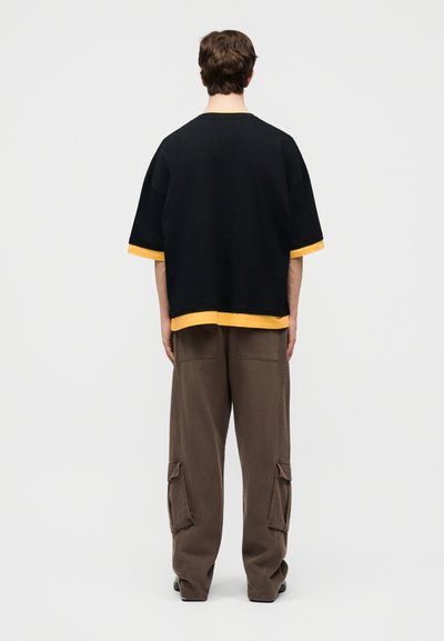 Mann steht mit dem Rücken zur Kamera, trägt ein schwarzes, übergroßes T-Shirt mit gelbem Rand und braune Cargohosen vor weißem Hintergrund.