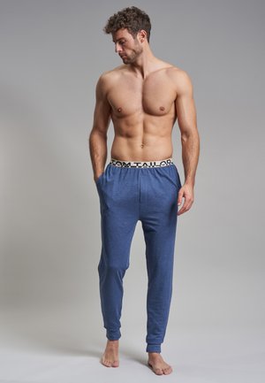Pantalon lounge en coton bleu avec taille élastique portant le logo "TOM TAILOR", jambes fuselées et poches latérales. Le modèle est pieds nus.