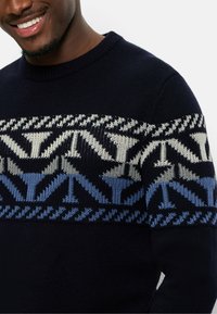 Marinblauer Strickpullover mit einem geometrischen Muster in Creme, Hellblau und Dunkelblau über der Brust, mit einem gerippten Rundhalsausschnitt.