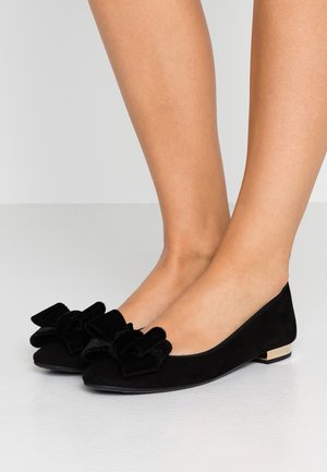 Zapatos planos de ante negro para mujer con grandes lazos de terciopelo en la parte superior y tacones bajos metálicos en bloque, usados con los pies descalzos sobre un fondo neutro.