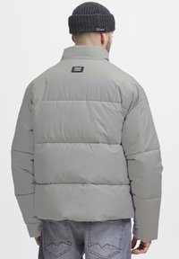 Gray puffer jacket with a high collar, featuring a thick quilted design, and a logo patch on the upper back. Worn with a knitted dark hat.