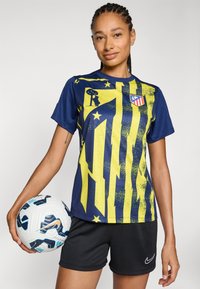 Nike nogometna majica u tamnoplavoj i žutoj boji, s prugastim dizajnom i grafičkim elementima, nosi se s crnim kratkim hlačama i drži loptu za nogomet.