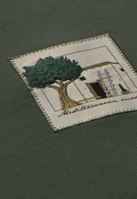 Tessuto verde con una toppa quadrata cucita, decorata con un albero ricamato e una casa, accompagnata dal testo "Umore mediterraneo."