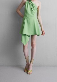 Grön halterneck-klänning med asymmetrisk fåll, tillverkad av lätt tyg. Matcher med glittrande, remmade gula klackar. Slät textur synlig.