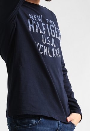 Navy blue long-sleeve t-shirt with white text “NEW YORK HILFGER U.S.A. MCMLXX” on front; cotton fabric, relaxed fit, round neckline.