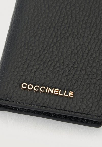 Coin de portefeuille en cuir noir texturé avec lettrage du logo en métal doré "COCCINELLE" sur fond clair.