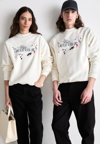 Deux personnes portent des sweats crème à col rond avec des motifs éclaboussés multicolores et du texte, associés à des pantalons noirs et un sac blanc.