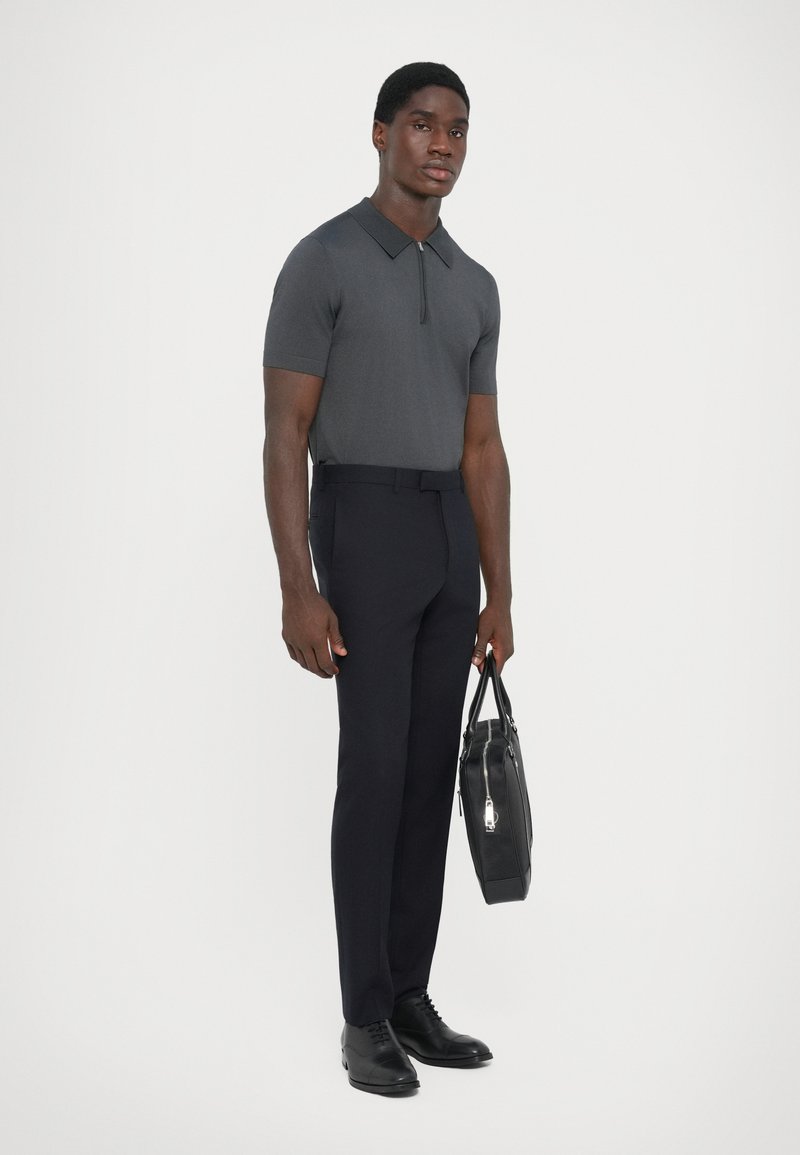 Graues Kurzarm-Polo mit Reißverschlusskragen, kombiniert mit schwarzen, maßgeschneiderten Hosen und schwarzen Lederschuhen. Die Person trägt eine schwarze Tasche.