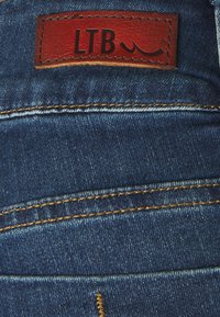 Vaqueros de denim de color azul medio, con un parche de cuero marrón etiquetado "LTB" y costuras amarillas a lo largo de las costuras. Textura suave.