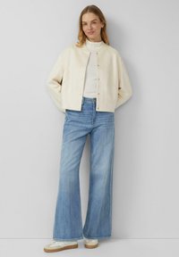 Cremefarbene, knopfgeknöpfte Jacke mit weiten Ärmeln, kombiniert mit hoch taillierten, hellblauen Schlagjeans und weißen Sneakers mit beigen Sohlen.