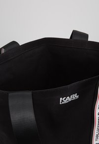 Svart Karl Lagerfeld tote-väska med svarta handtag och vit logotyp tryckt nära övre kanten, som visar ett tomt inre mot en grå bakgrund.