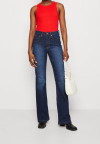 Hoge taille flared jeans in donkerblauwe denim, gecombineerd met een aansluitend rood geribbeld tanktop. Model houdt een witte gequiltte handtas vast.