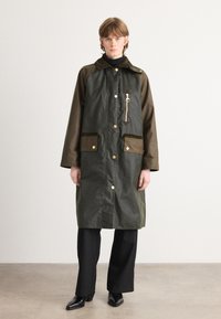 Groene waterproof jas met bruine accenten, twee voorzakken, drukknoopsluitingen en een ritsdetail. Draagt over een zwarte coltrui en broek.
