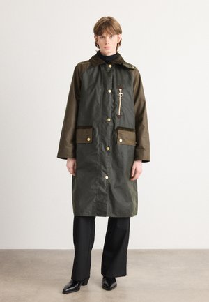 Groene waterproof jas met bruine accenten, twee voorzakken, drukknoopsluitingen en een ritsdetail. Draagt over een zwarte coltrui en broek.