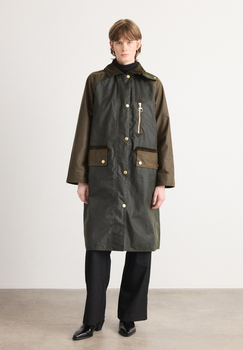 Groene waterproof jas met bruine accenten, twee voorzakken, drukknoopsluitingen en een ritsdetail. Draagt over een zwarte coltrui en broek.