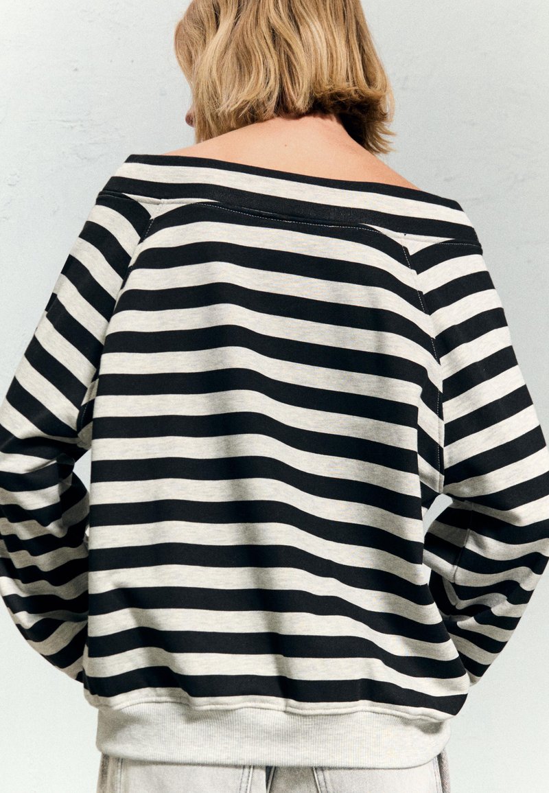 Osoba z krótkimi blond włosami nosząca bluzkę z długim rękawem odsłaniającą ramiona, w czarno-białe poziome paski, odwrócona plecami.