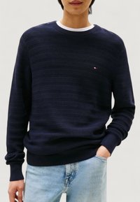 En person som bär en marinblå strukturerad tröja med rund halsringning och en liten logotyp på bröstet, samt ljusblå jeans, med en hand i fickan, på en neutral bakgrund.