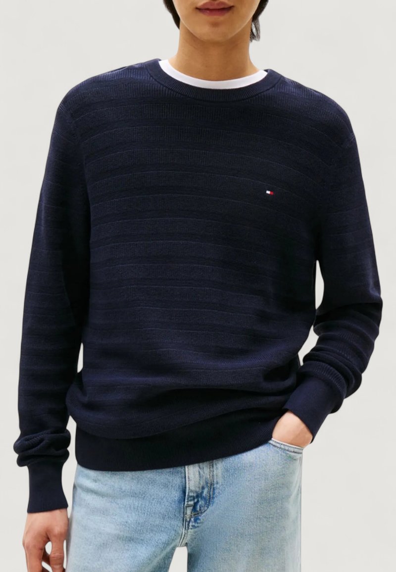 En person som bär en marinblå strukturerad tröja med rund halsringning och en liten logotyp på bröstet, samt ljusblå jeans, med en hand i fickan, på en neutral bakgrund.