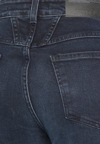Tmavě modré džíny z denimu s přiléhavým střihem, které vystihují jemné textury, standardní šití a dvě zadní kapsy. Na pase je viditelná značka.
