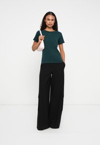 Tricou scurt de culoare verde închis, cu guler rotund, asortat cu pantaloni lărgi negri, prevăzuți cu buzunare laterale. Modelul ține o geantă mică roz.
