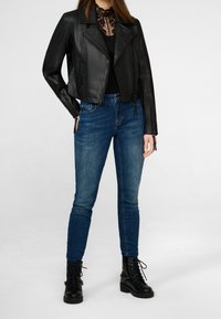 Chaqueta corta de cuero negro con cierre frontal, combinada con jeans skinny azules y botas de tobillo negras con cordones. Cuello plano y textura detallada.