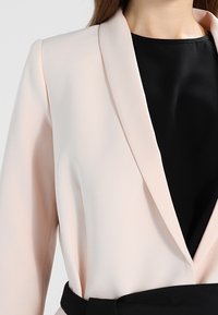 Jasnoróżowa marynarka z kołnierzem szalowym, długimi rękawami i elegancką czarną podszewką. Posiada pasek w talii oraz miękki, gładki materiał.