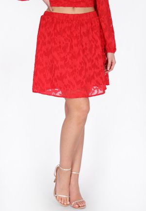 Gonna rossa testurizzata al ginocchio abbinata a top corto coordinato e décolleté nude con cinturini alla caviglia su sfondo bianco.