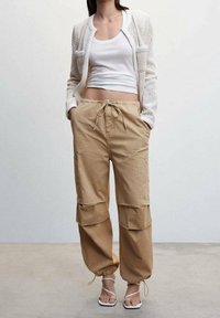 Pantalons cargo beiges avec des poignets élastiques, des poches latérales et une taille à cordon, associés à un débardeur blanc court et un cardigan blanc texturé.