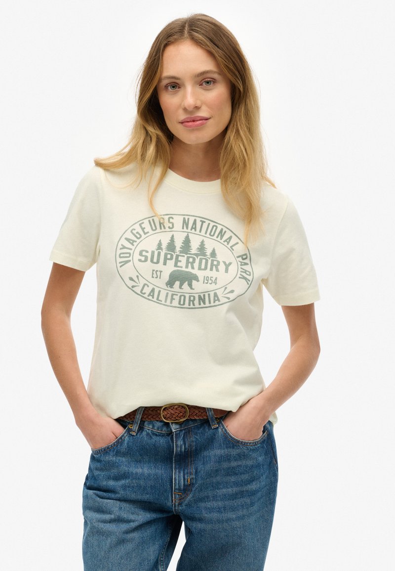 Kremowa koszulka z nadrukiem przedstawiającym drzewa i niedźwiedzia, połączona z niebieskimi spodniami o wysokim stanie i brązowym plecionym paskiem.