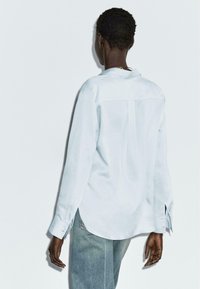 Camicia a maniche lunghe azzurro chiaro in tessuto morbido, con un dettaglio di yoke posteriore e fessure laterali vicino all'orlo. Indossata con jeans in denim chiaro.