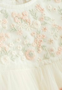 Tissu blanc avec une broderie florale douce dans des teintes roses et vertes, présentant des coutures délicates et un ourlet volanté pour ajouter de la texture.