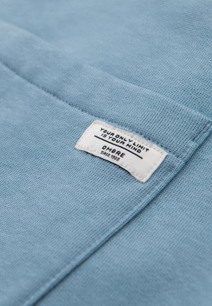 Lyst blå bomuldsstof med en tekstureret overflade, der har et hvidt mærke prægede med "DIN ENESTE GRÆNSE ER DIN HJERNE" og "OMBRE Siden 1985."